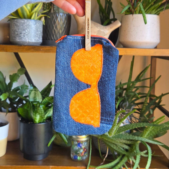 Hand holding a small, handcrafted denim sunglass pouch with orange accents against a plant-filled background