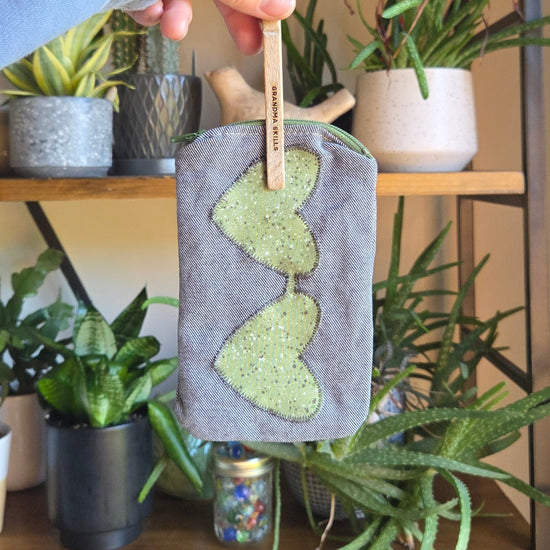 Hand holding a small, handcrafted denim sunglass pouch with green accents against a plant-filled background