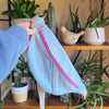 Light blue denim fanny pack with a mauve zipper held by a person in front of a decorative background with plants and shelves.