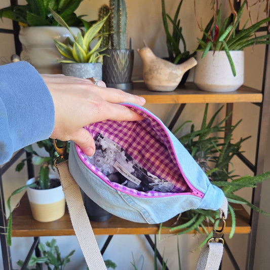 Light blue denim fanny pack with a mauve zipper held by a person in front of a decorative background with plants and shelves.